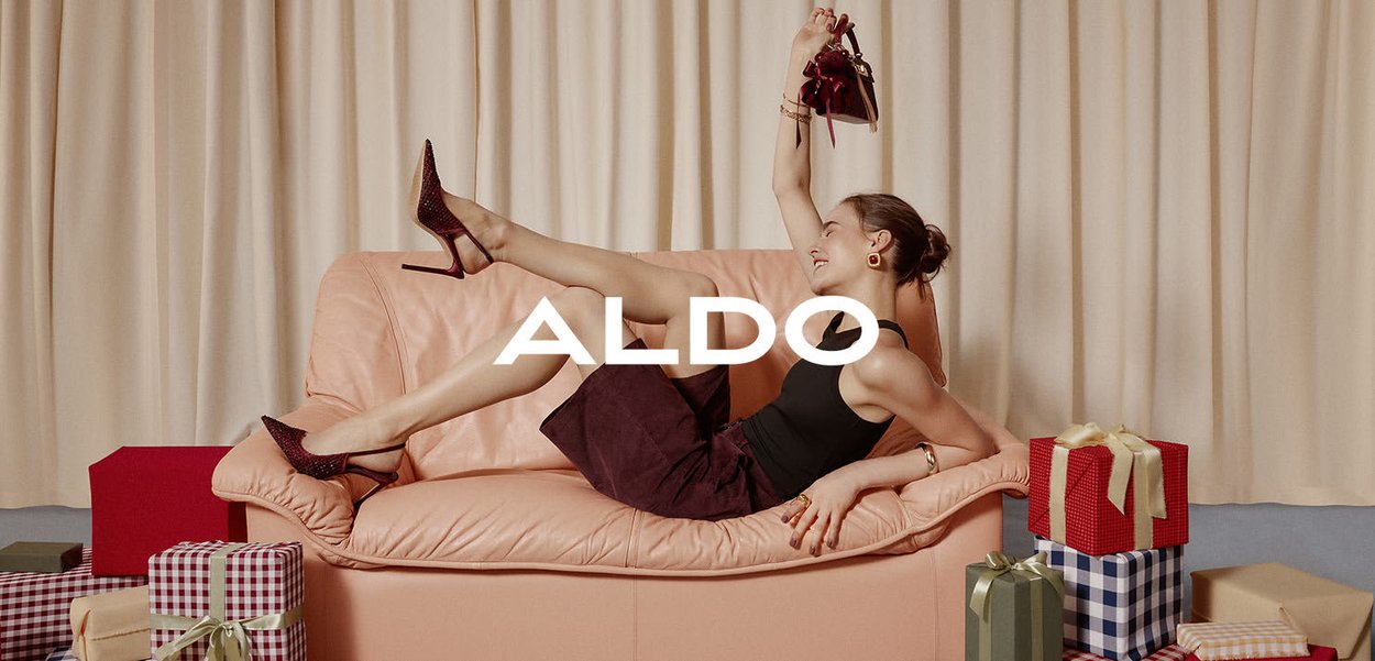 Roze leren bank met een model in een zwarte tanktop en een bordeauxrood rokje, die rode schoenen en een handtas vasthoudt, omringd door kleurrijke geschenkdozen.