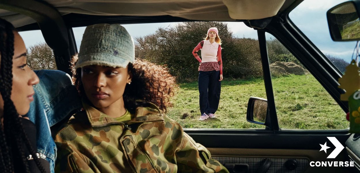Dos personas están sentadas dentro de un coche, una con ropa de camuflaje, mientras que una tercera persona está de pie afuera sobre el césped, vistiendo ropa rosa y azul marino, bajo un cielo despejado.