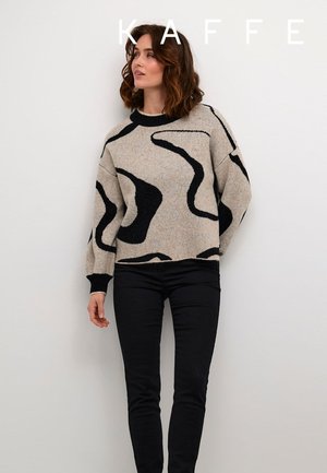 Beige neulepaita mustalla abstraktilla kuviolla, pyöreä pääntie ja laskeutuvat olkapäät. Yhdistetty mustiin housuihin, seisoo valkoista seinää vasten.