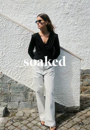 Schwarzes Langarmoberteil mit Wasserfallausschnitt, kombiniert mit hoch taillierter weißer weiter Hose. Im Hintergrund eine strukturierte weiße Wand und Steinpflaster.