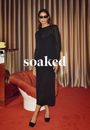 Robe en tricot noir à côtes avec manches longues, longueur cheville et silhouette ajustée. Modèle portant des lunettes de soleil noires. Chaise orange vif en arrière-plan.