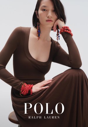 Vrouw in een bruine jurk met lange mouwen zit, draagt grote rode en blauwe kralen oorbellen en meerdere rode kralen armbanden, het hoofd leunt op de hand.