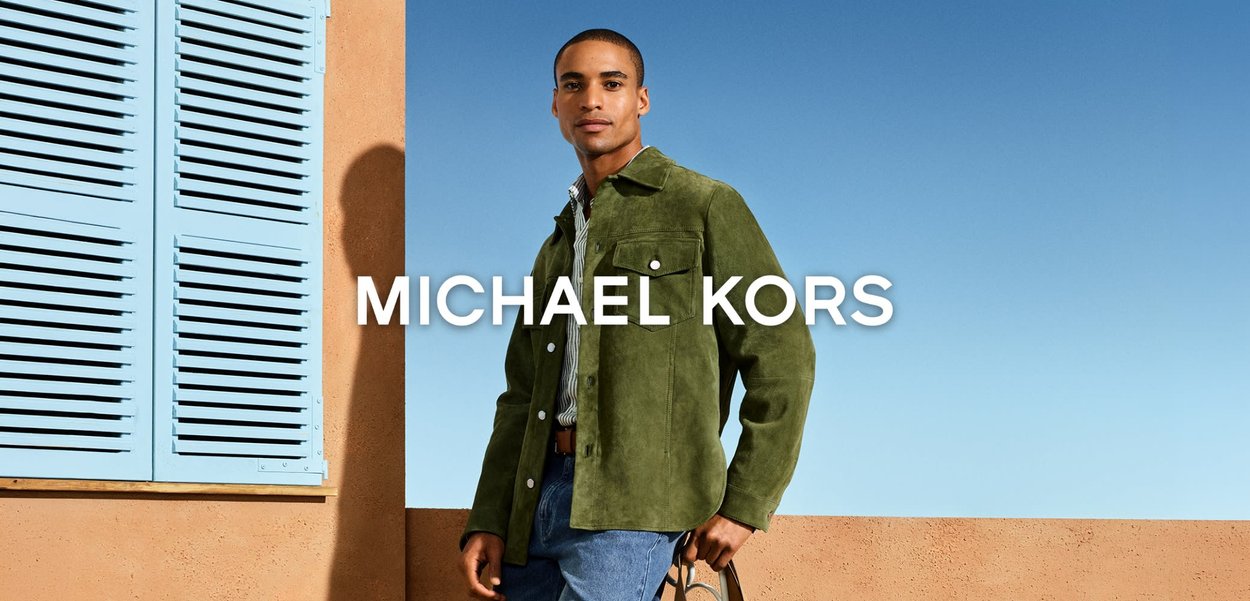 Hombre con chaqueta de ante verde y vaqueros azules de pie al aire libre frente a una pared de terracota y una ventana con contraventanas azules bajo un cielo despejado.