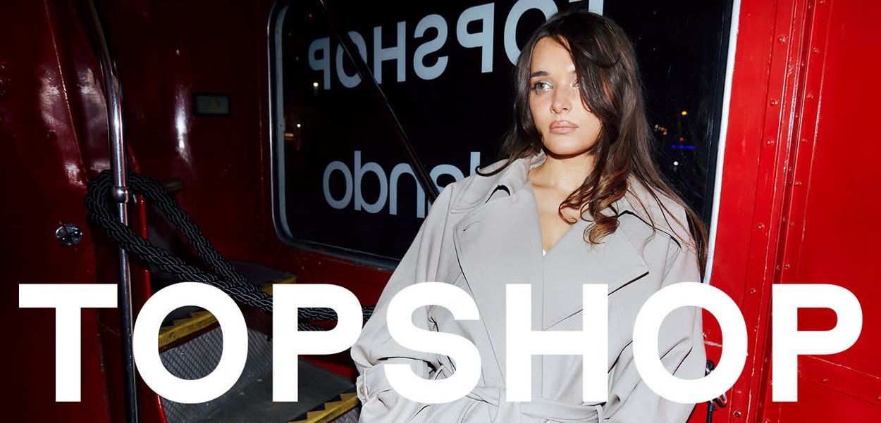 Junge Frau in einem beigen Trenchcoat sitzt in einem roten Bus, auf dem Bild ist der Text "TOPSHOP" eingeblendet.