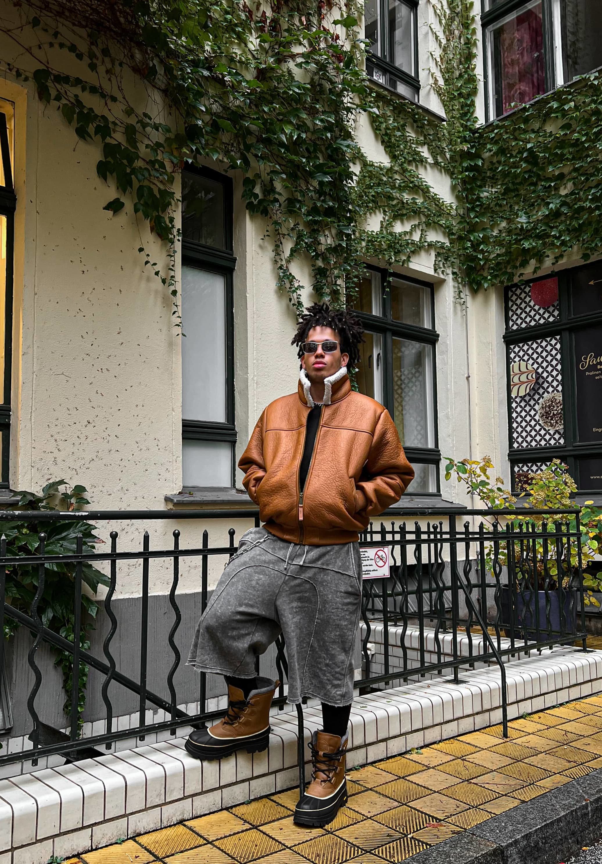 Veste en cuir marron type bombardier avec doublure blanche, shorts gris surdimensionnés et bottes à lacets marron sur un chemin carrelé avec de la verdure en arrière-plan.