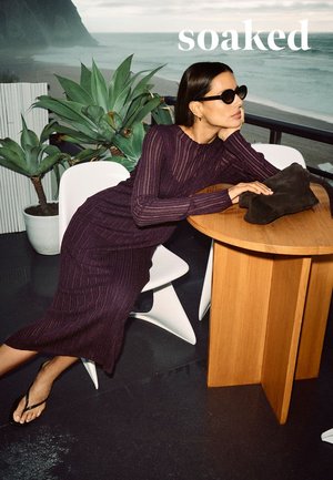 Vestito viola in maglia a coste, maniche lunghe, tessuto strutturato. Borsa nera su tavolo di legno. Sedia moderna bianca, piante verdi sullo sfondo.