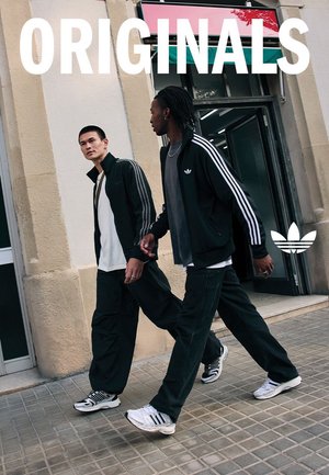 Due persone camminano fianco a fianco indossando tute nere con strisce bianche. Portano scarpe da ginnastica e si trovano su una superficie piastrellata fuori da un negozio.