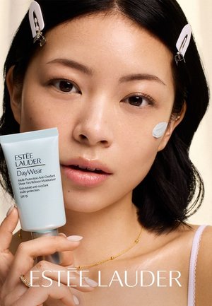 Ung kvinde med kort mørkt hår og clips holder Estée Lauder DayWear fugtighedscreme, med en lille mængde påført på hendes kind.