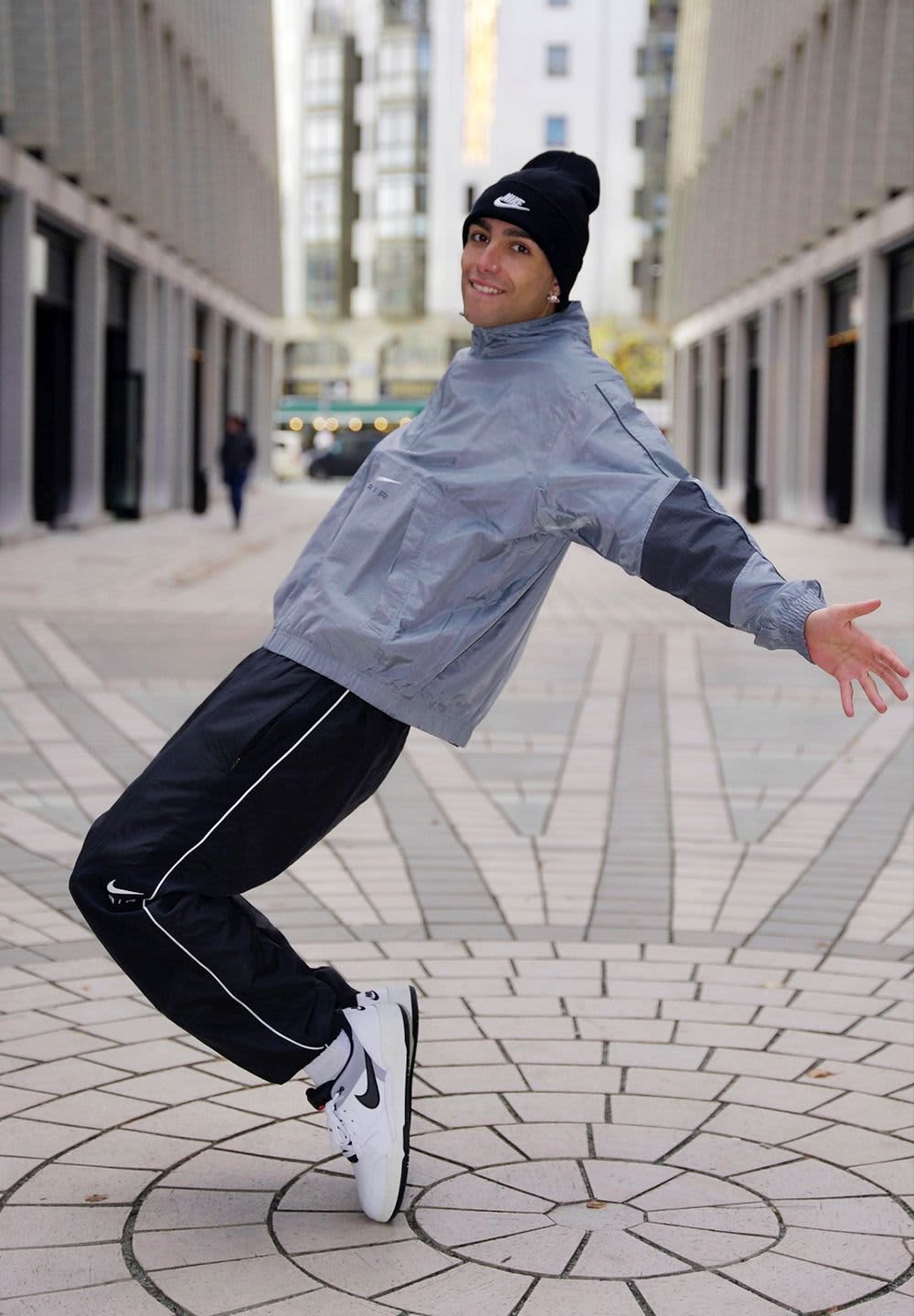 Athlete in gray windbreaker with black accents and matching black track pants with white stripes, wearing black beanie and white sneakers.