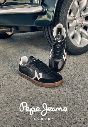 Zapatillas Pepe Jeans en blanco y negro con suelas marrones sobre asfalto cerca de la llanta de un coche, con el logo de Pepe Jeans London debajo.