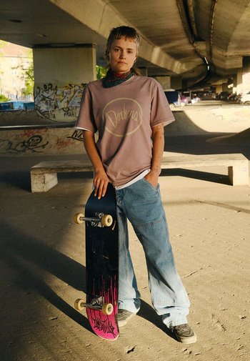 T-shirt graphique violet avec le logo "Dockers", jean bleu décontracté, skateboard noir avec des accents roses et baskets grises. Cadre d'un skatepark en plein air.