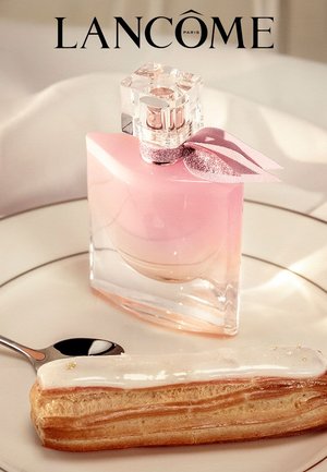 Rosa Lancôme Parfümflasche mit einem klaren Glasboden und einem Kristallverschluss, neben einer glasierten Süßspeise auf einem weißen Teller mit einem silbernen Löffel.