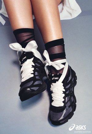 Piernas con calcetines negros transparentes y zapatillas negras Asics con cordones de cinta blanca de satén de gran tamaño atados en lazos sobre un suelo gris.