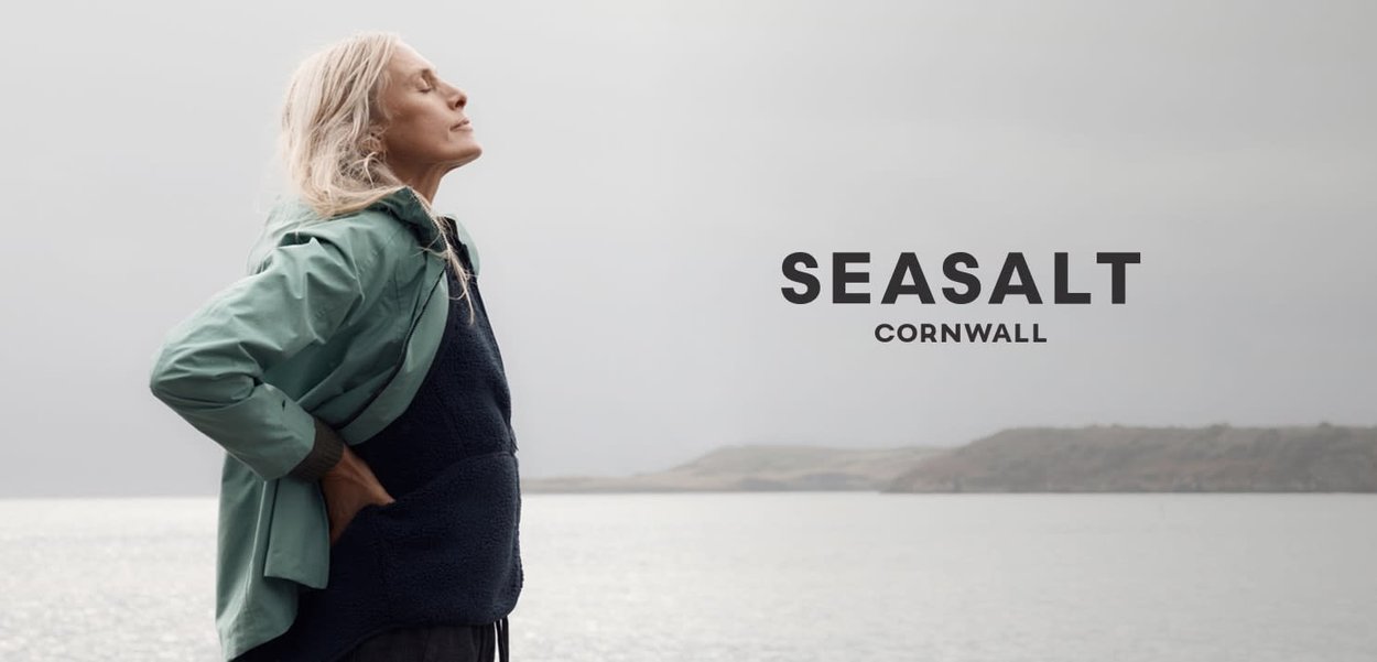 Femme âgée aux cheveux blancs debout près d'une mer calme, vêtue de plusieurs couches de vêtements, les yeux fermés, face à la brise et à la côte lointaine sous un ciel gris.