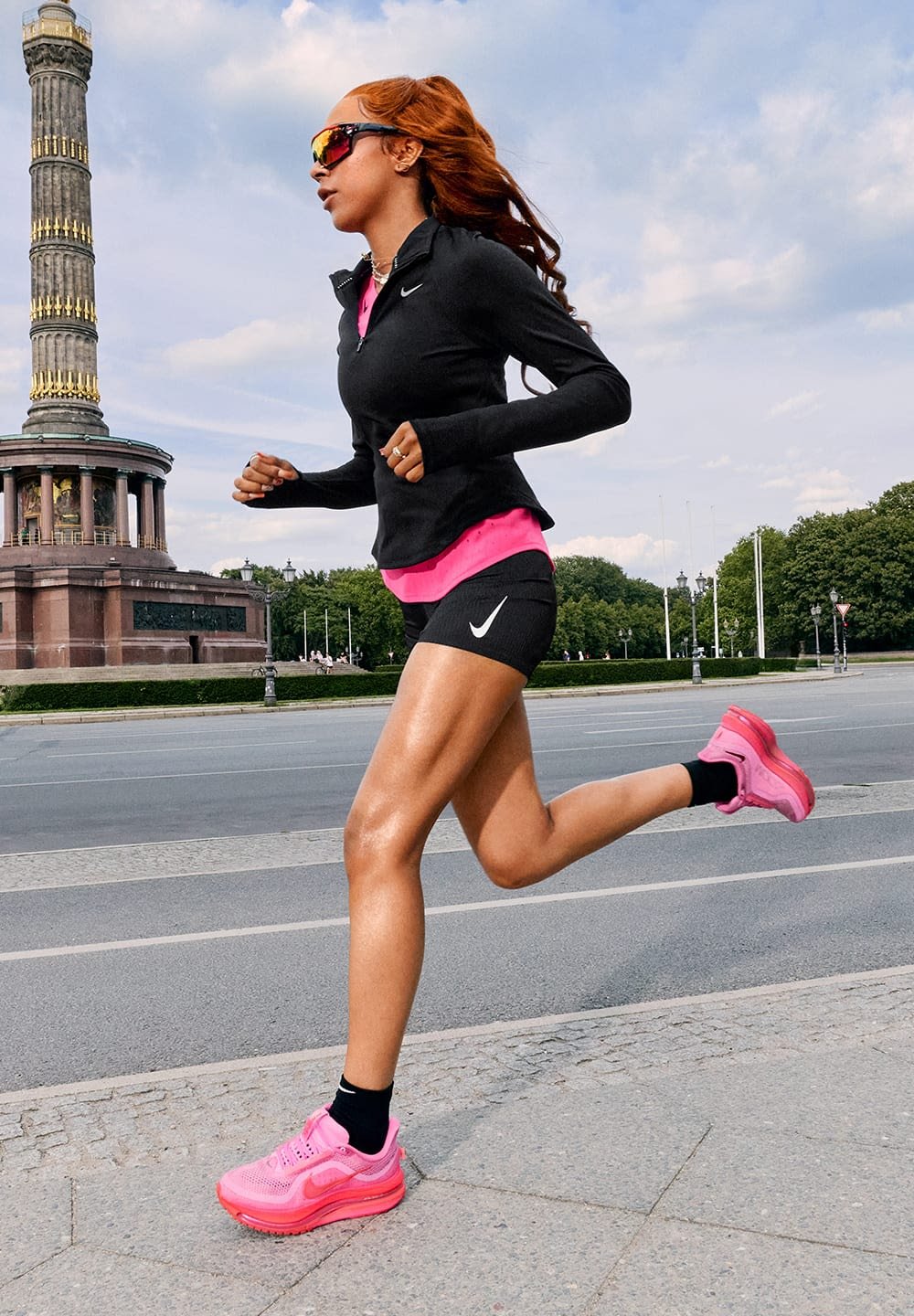 Top nero a maniche lunghe, camicia rosa, pantaloni corti neri e scarpe da corsa rosa. Corridore in movimento, corporatura atletica, con occhiali da sole, sfondo urbano.