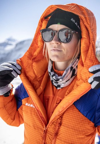 Frau stellt die Kapuze ihrer orange-blauen Winterjacke ein, trägt eine schwarze Mütze, Sonnenbrille, Handschuhe und einen Halswärmer in einer schneebedeckten Berglandschaft.