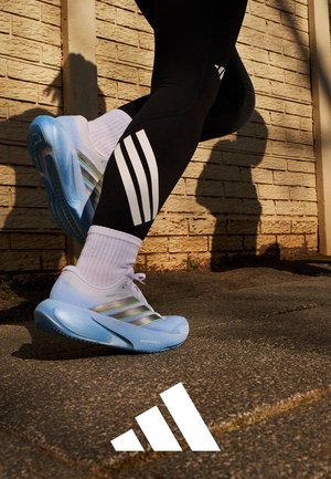 Løper iført svarte leggings med hvite striper og hvite Adidas-sneakers med blå såler, går på fortauet nær en murvegg.