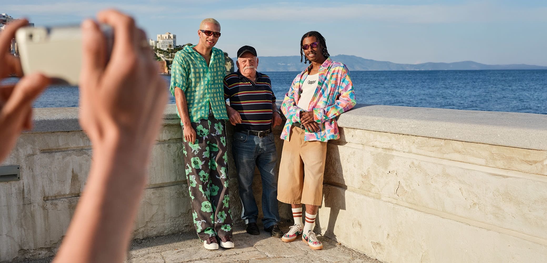 Troje pojedinaca stoje na kamenoj ogradi uz more. Jedan nosi košulju s zelenim uzorkom i crne cvjetne hlače, drugi prugastu košulju, a treći šareni košulju s dugmićima i bež šorts.
