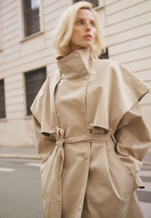 Blonde Frau trägt einen beigen Trenchcoat mit hohem Kragen und Cape-Detail, Hände in den Taschen, steht auf einer Stadtstraße.