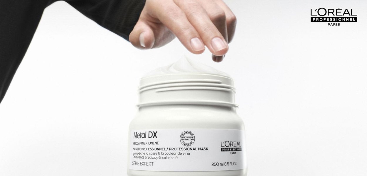 En hånd strækker sig mod et åbent hvidt glas af L’Oréal Metal DX Professionel Maske med flødeskum svunget ovenpå, på en hvid baggrund.