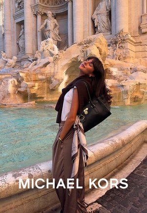 Borsa nera Michael Kors con accessori in argento. Il modello indossa un top bianco con un senza maniche marrone e pantaloni beige. Fontana di Trevi sullo sfondo.