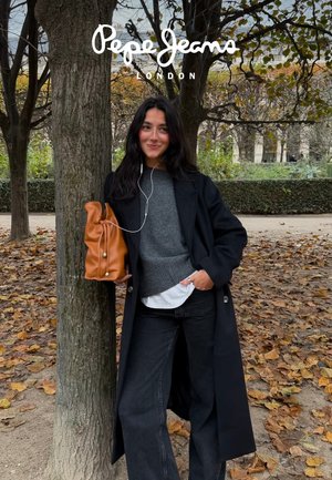 Een vrouw leunt tegen een boom, draagt een lange zwarte jas, een grijze trui en donkere spijkerbroek, en houdt een bruine tas met een ontspannen ontwerp vast.