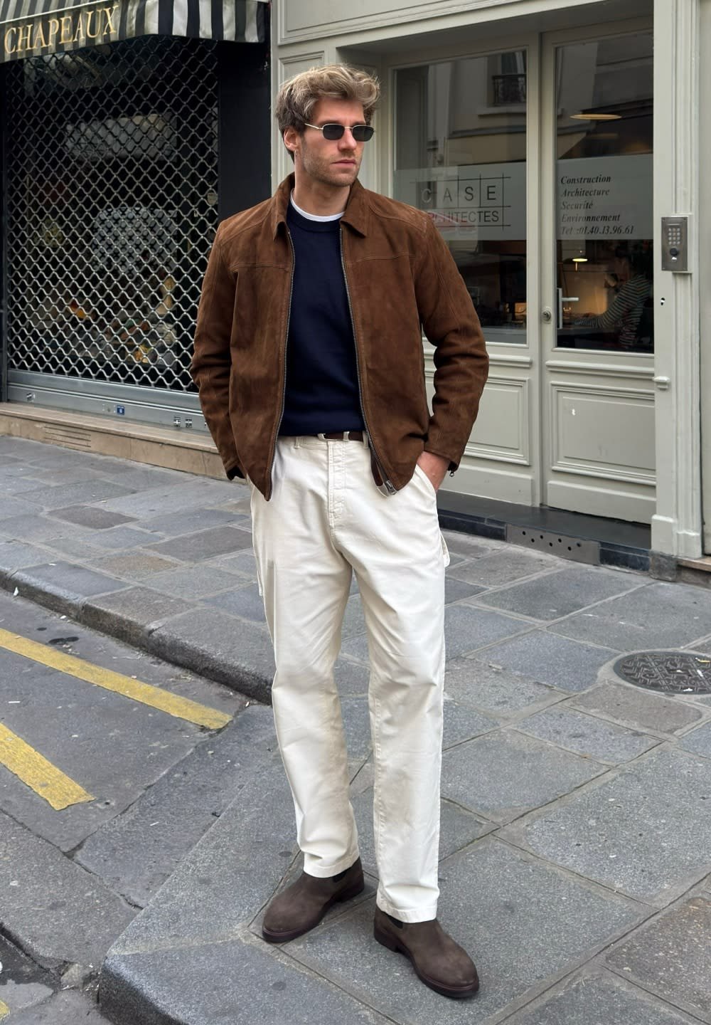 Bruine suède jas over een marineblauw shirt, crèmekleurige broek en bruine laarzen. Zonnebril en kort, gestructureerd haar. Stedelijke omgeving met winkels.