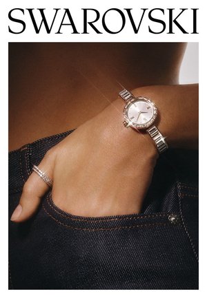 Orologio da polso Swarovski con bracciale in metallo di tonalità argentata, quadrante rotondo bianco e dettagli in cristallo. Indossato con un anello incastonato di diamanti, su sfondo di denim scuro.