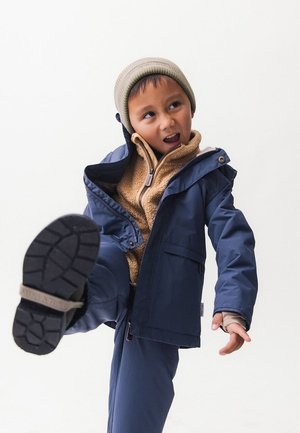Navy waterproof jacket with hood over a tan fleece sweater. Gray beanie, gray gloves, and black rubber-soled shoes with a tread pattern.