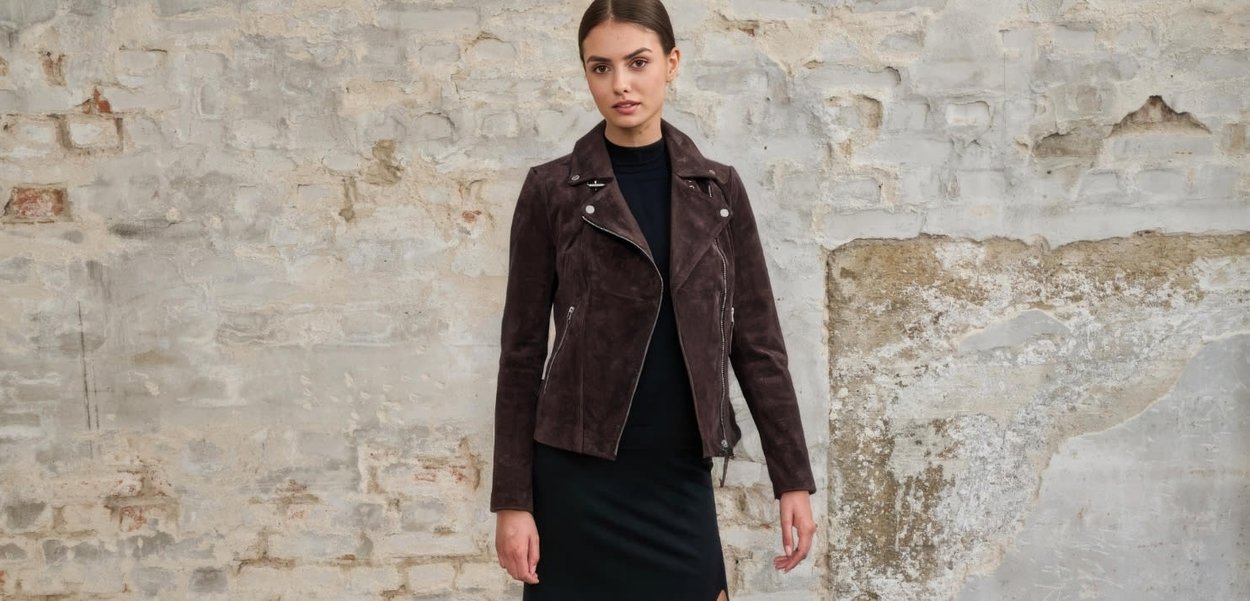 Kobieta w ciemnej zamszowej kurtce motocyklowej na czarnej sukience stoi na tle teksturowanej jasnoszarej ściany z cegieł.