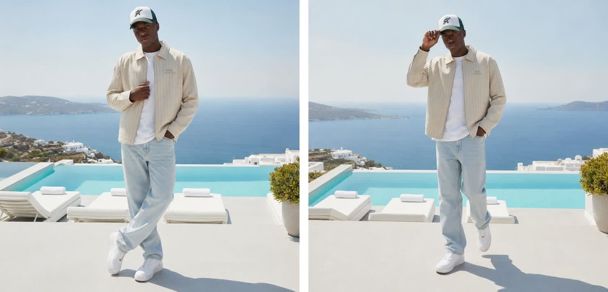 Mann mit leichter Jacke, weißem Hemd, Jeans und Kappe posiert nahe dem Pool mit Ozean und Hügeln im Hintergrund unter klarem Himmel.