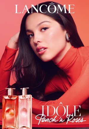 Femme en col roulé orange appuyée sur une table à côté de deux flacons de parfum Lancôme Idôle de couleur pêche et rose, sur un fond pêche.