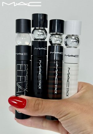 Four MAC Stack mascara tubes in varying colors: black, dark brown, and white, with rounded caps and transparent spherical accents.