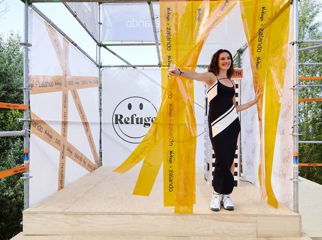 Schwarz-weißes Kleid mit asymmetrischen Streifen, umgeben von gelben durchsichtigen Vorhängen in einer holzverkleideten Struktur mit Branding-Elementen.