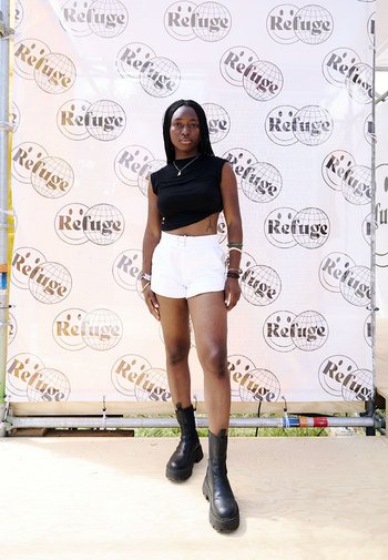 Schwarzes kurzes T-Shirt, weiße hoch taillierte Shorts und schwarze Ankle Boots. Steht vor einem markierten Hintergrund mit wiederholten Logos.