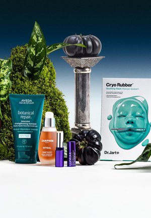 Bottle of green hair mask, orange serum, and two purple serums, with a teal facial mask box and dark vegetables among plants on white background.