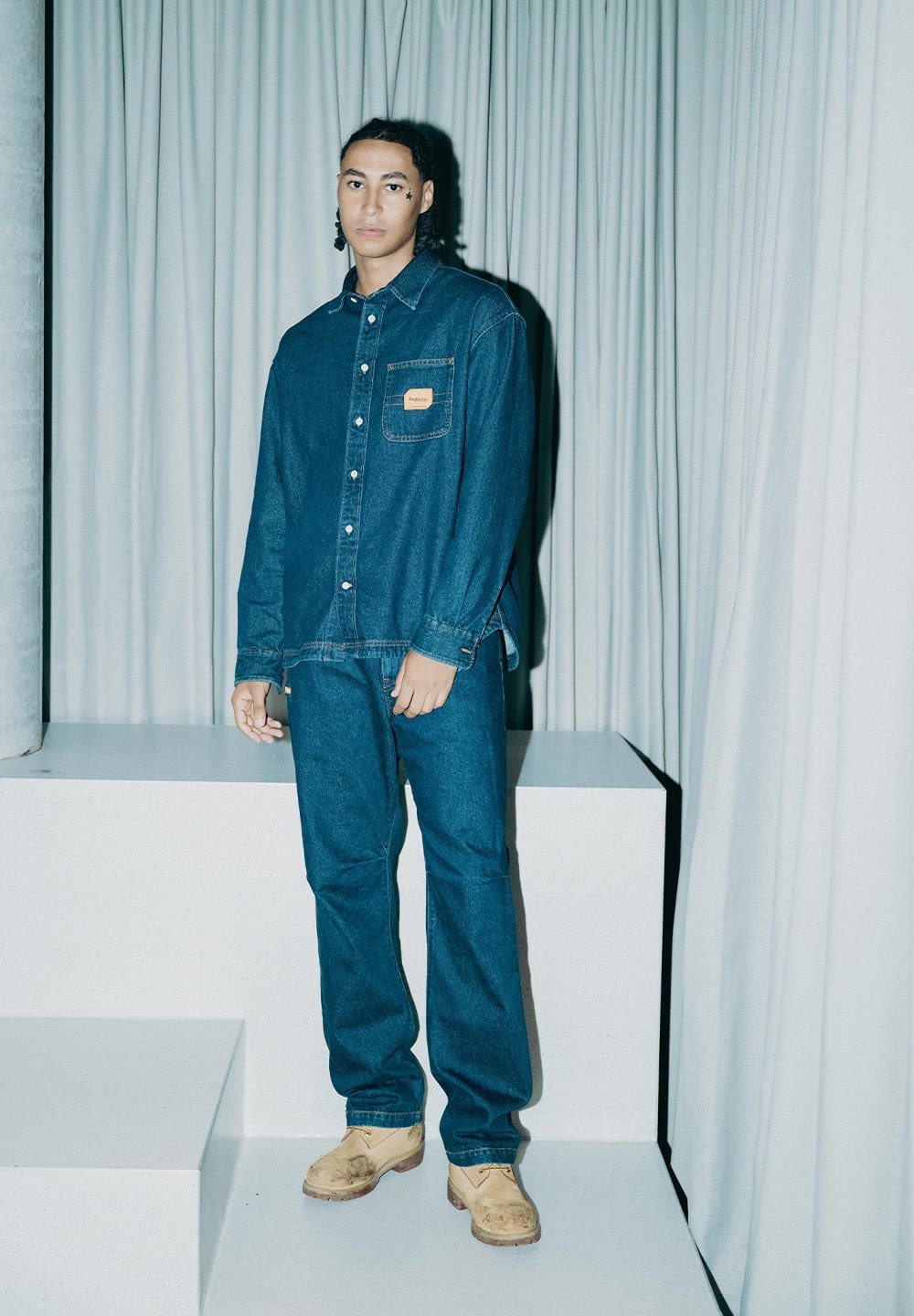 Junge Person mit geflochtenem Haar, die ein blaues Denim-Hemd und eine Jeanshose mit beigen Schuhen trägt, steht vor weißen Vorhängen und geometrischen weißen Strukturen.