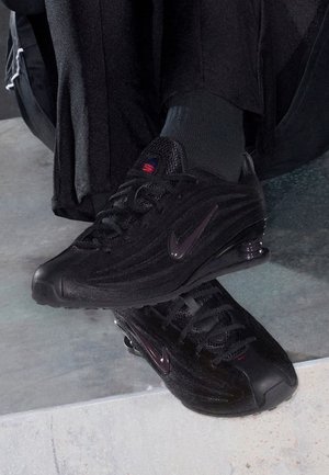 Persona con zapatillas negras Nike y calcetines oscuros, apoyando un pie sobre una superficie de cemento en interiores.