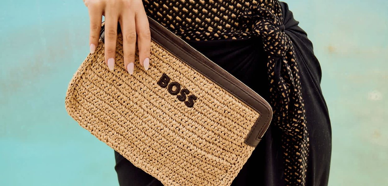 Mano sosteniendo un bolso de mano tejido de color marrón con el logo "BOSS", en contraste con una tela negra y un fondo turquesa.