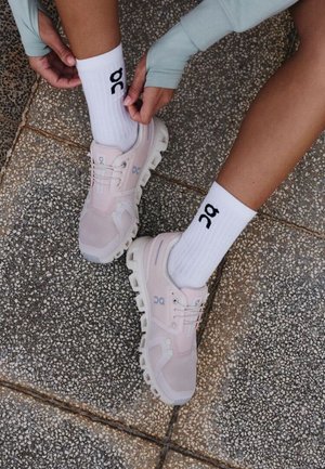 Person trägt blassrosa Laufschuhe und weiße Socken mit dem "OC"-Logo und zieht die Socke hoch, während sie auf einem gefliesten Gehweg sitzt.