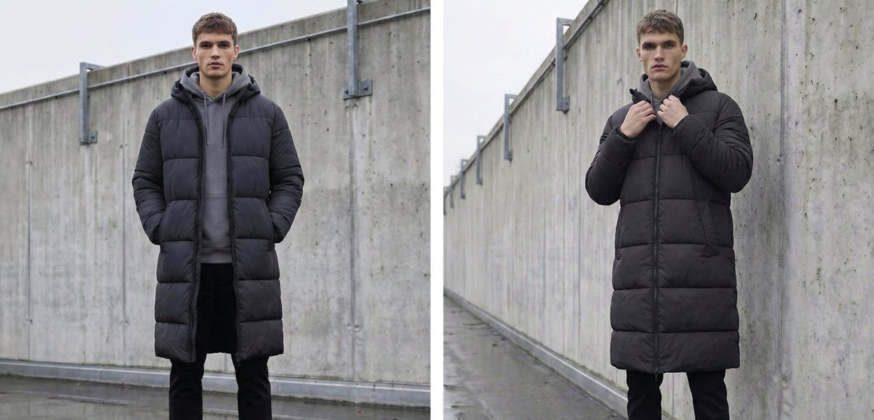 Giovane uomo in piedi contro un muro di cemento che indossa un cappotto imbottito lungo nero, una felpa con cappuccio grigia e pantaloni neri all'aperto.