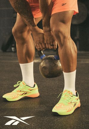 Persoon tilt een kettlebell op, draagt neongele en oranje Reebok-schoenen, witte sokken en oranje shorts, op een gymvloer.
