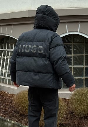 Giacca imbottita nera con cappuccio, caratterizzata da una texture opaca e dalla scritta "HUGO" in un font grande e audace sul retro.