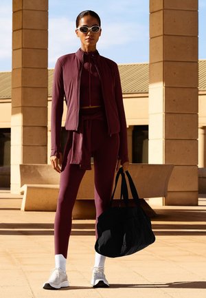 Maroon athletic outfit featuring a zip-up jacket and leggings. Accessories include black tote bag and white sneakers. Smooth texture, fitted design.