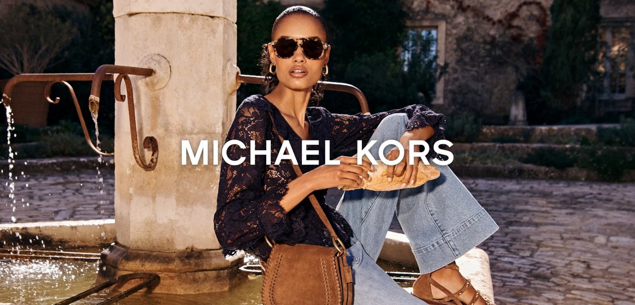 Kvinne med solbriller iført svart bluse av blonder og jeans sitter ved en steinfontene med brød i hånden, med brun skinnveske og sandaler.