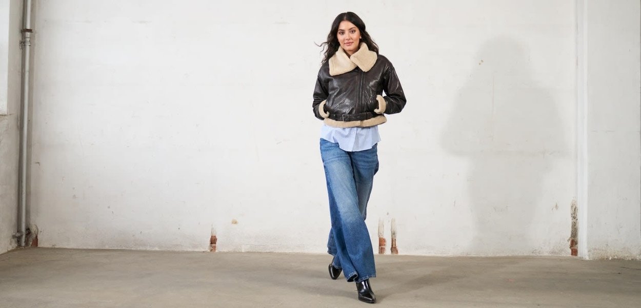 Giacca aviator in pelle marrone con colletto e polsini in pelliccia beige, indossata sopra una camicia a righe blu, abbinata a jeans a gamba larga blu e tacchi neri.