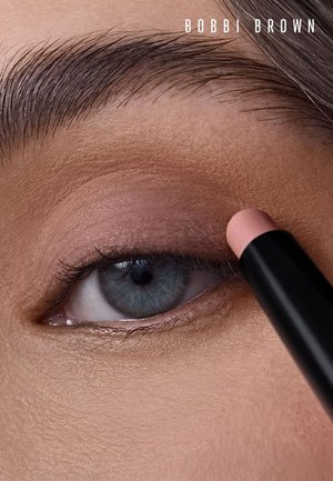 Gros plan sur un œil bleu avec des sourcils naturels, appliquant un fard à paupières rose doux à l'aide d'un stick de maquillage de la marque Bobbi Brown.