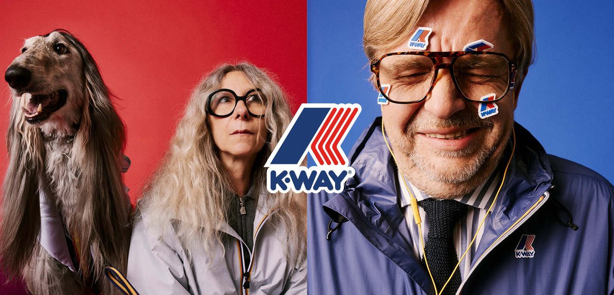 Chaquetas K-Way en púrpura y gris con cremalleras, llevadas por una persona con gafas y un galgo afgano. Coloridos adhesivos de K-Way adornan la cara del hombre.