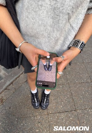 Chaussures de sport noires au design élégant et à la surface texturée, portées avec des chaussettes blanches. Un smartphone est tenu, affichant une image des chaussures.