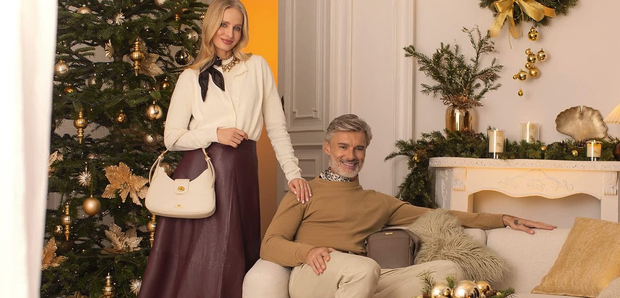 Dwie modelki siedzą, prezentując jasną torebkę, brązowy sweter i bordową spódnicę, na tle udekorowanego świątecznego dekoru.